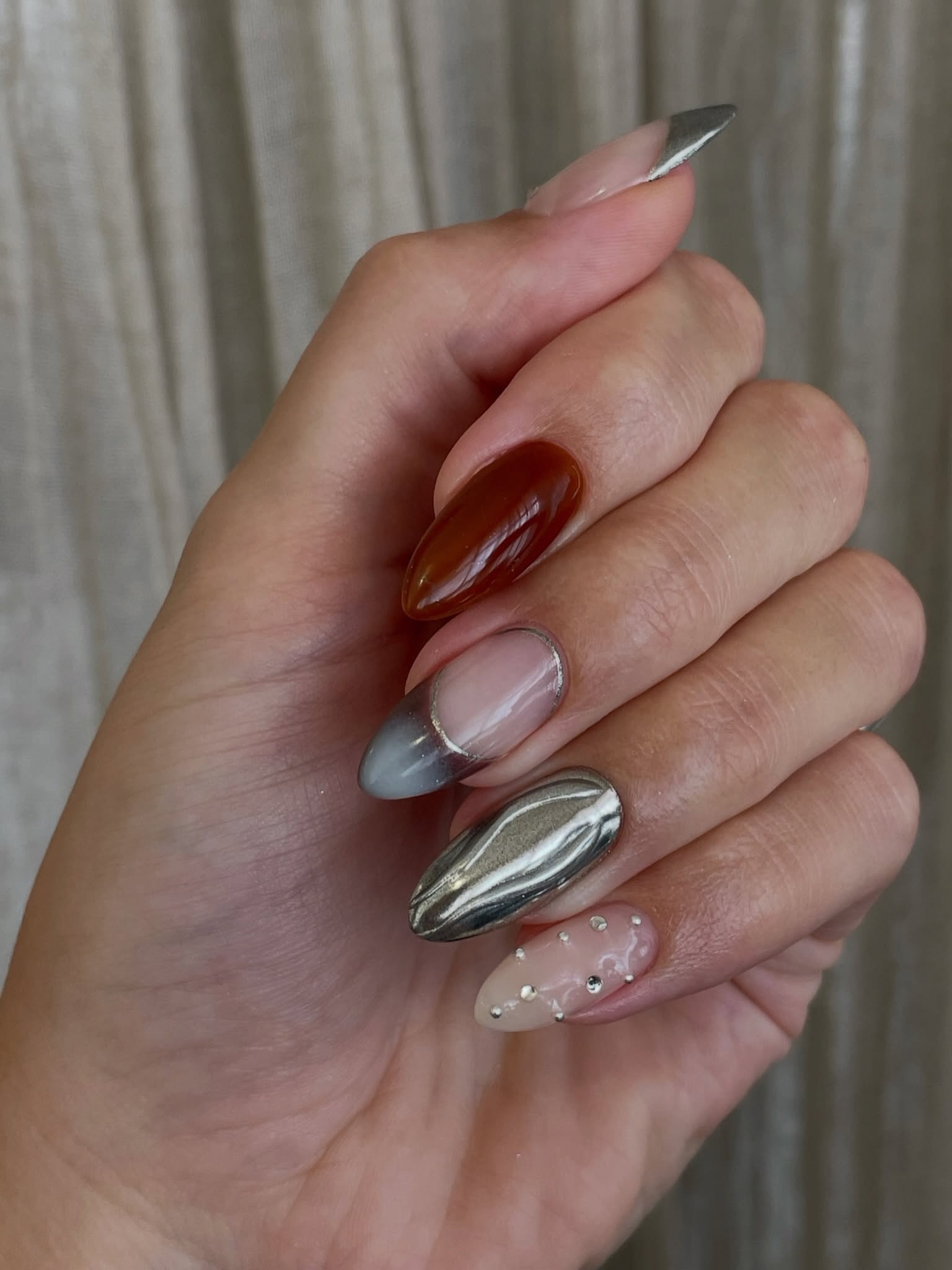 Mismatched almond nails with chocolate brown, silver chrome, sheer nude polish, and tiny gem details