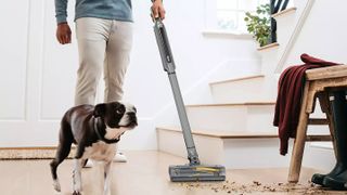 A man using the Shark WandVac to clean up spills ;on a wooden floor next to some stairs with a dog beside him