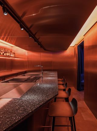 A orange-tinted room infused with warm light features a stone and stainless steal bar counter, matching chairs, and plenty of spirits on display.