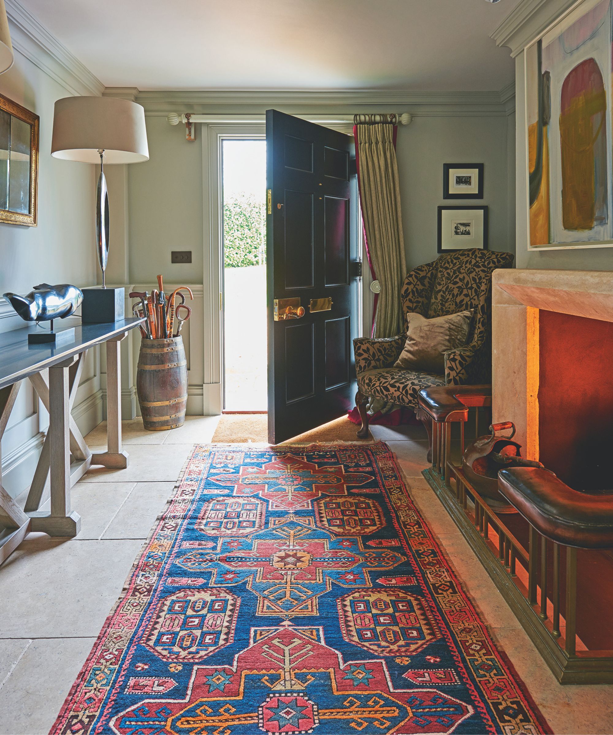 A cozy entryway with tiled flooring, a patterned blue and red rug, and a lit stone fireplace to the left with a large colorful artwork above. The dark blue front door to the house is open, with an umbrella stand filled with umbrellas, a white lamp, and side table to the left. On top of the table is a statue of a fish, with an artwork above.