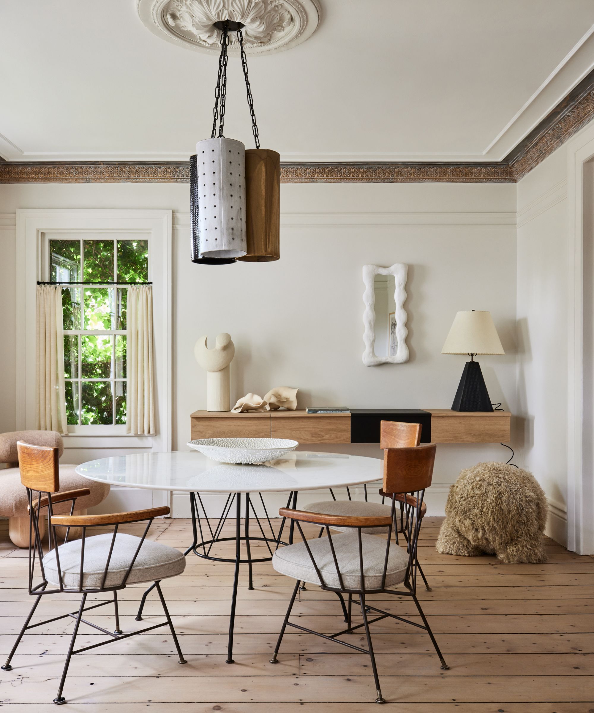 An airy dining area featuring a white marble table with wire-frame chairs, set against a minimalist console and a decorative floral ceiling medallion