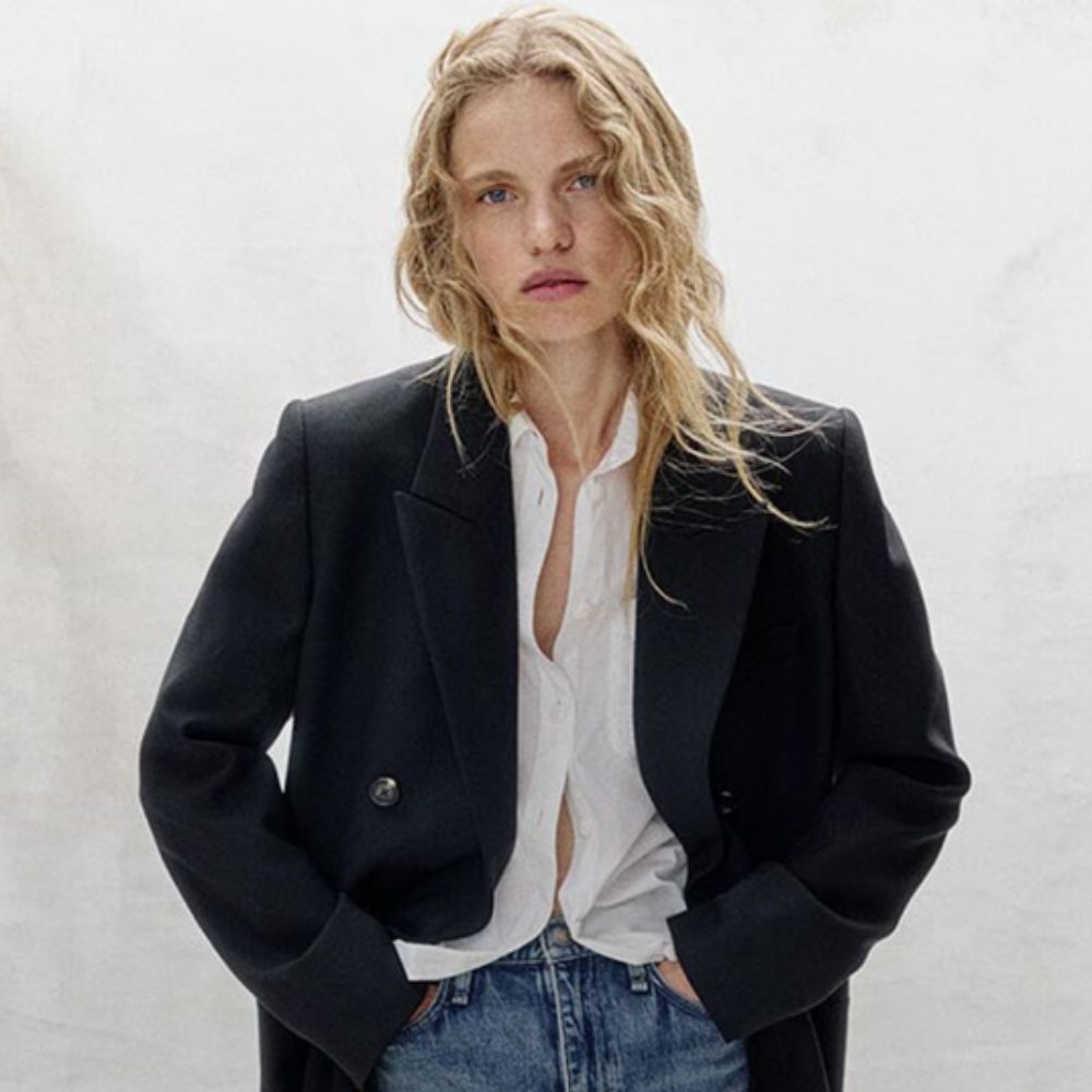 Blonde woman staring at camera wearing jeans, white blouse and dark coat. 