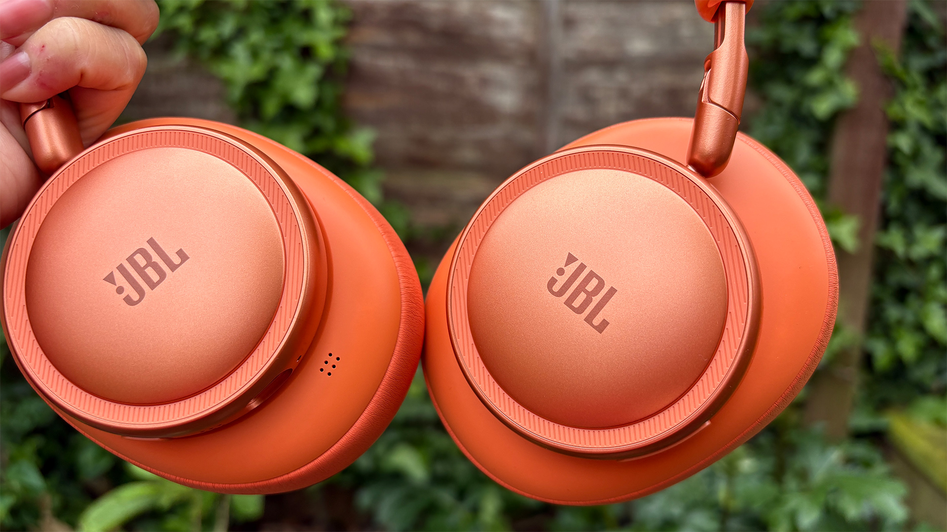 JBL Live 780NC wireless headphones held in hand in front of fence showing JBL logos on outer earcups