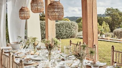 Three hanging rattan lampshades above a table set for outdoor dining, view of garden