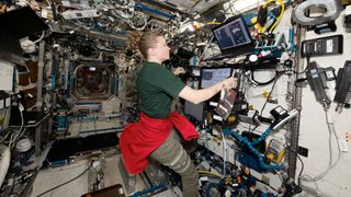 a woman in a forest green short sleeve shirt, kahki slacks and a red sweatshirt tied around her waist works with several laptop computers inside a space station module