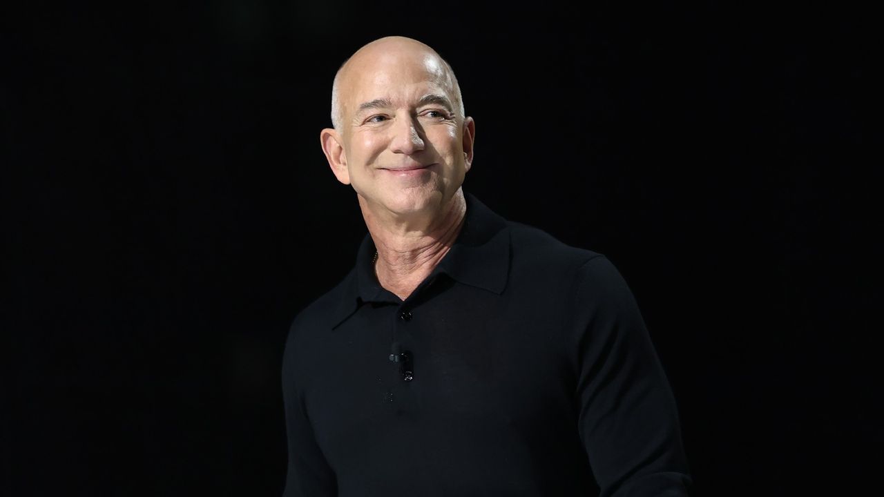 front shot of Jeff Bezos, dressed in a black pullover in front of a black background. Bezos is smiling lightly