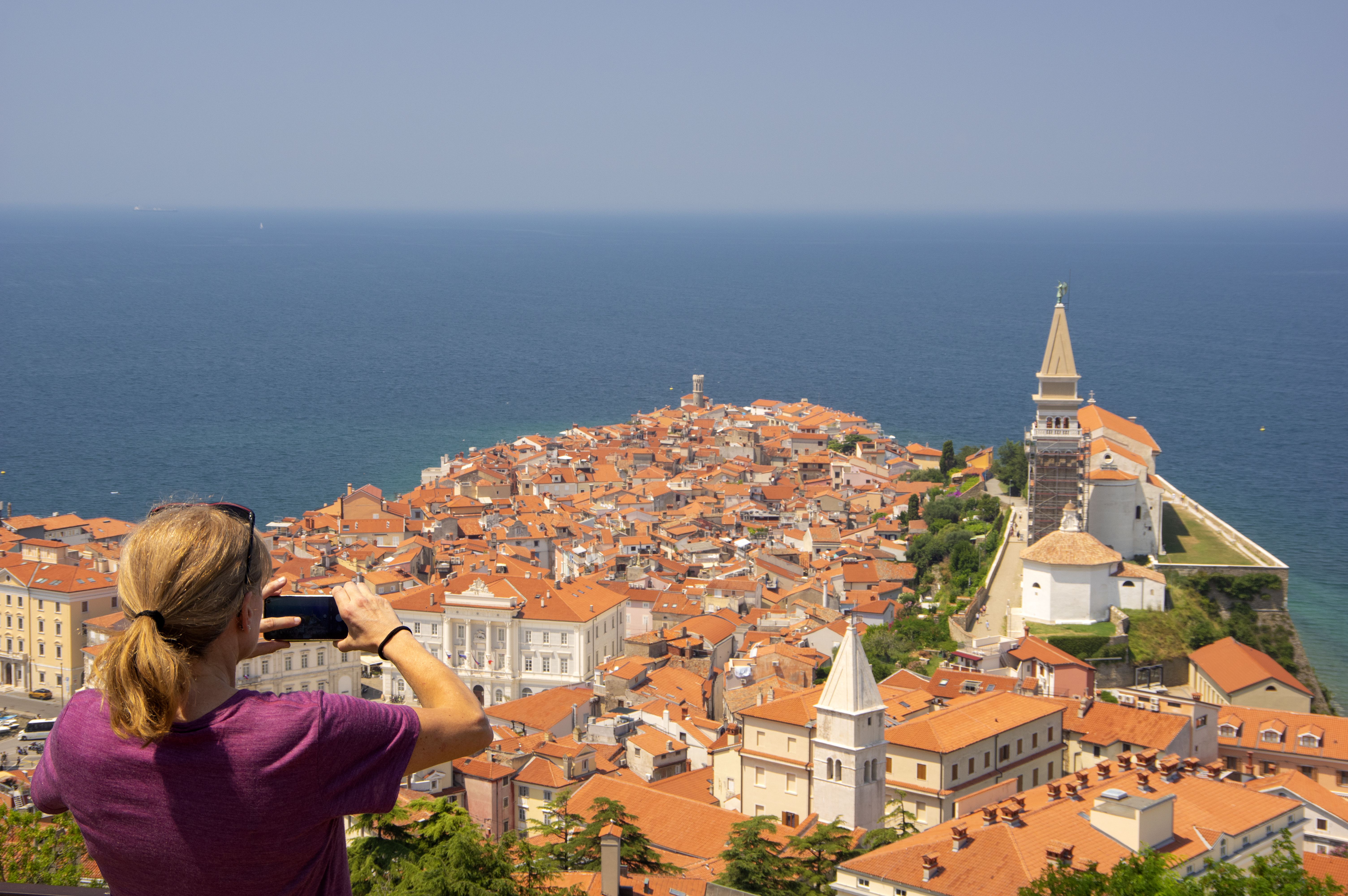 A woman wearing a purple shirt takes a photo of the town of Piran, Slovenia