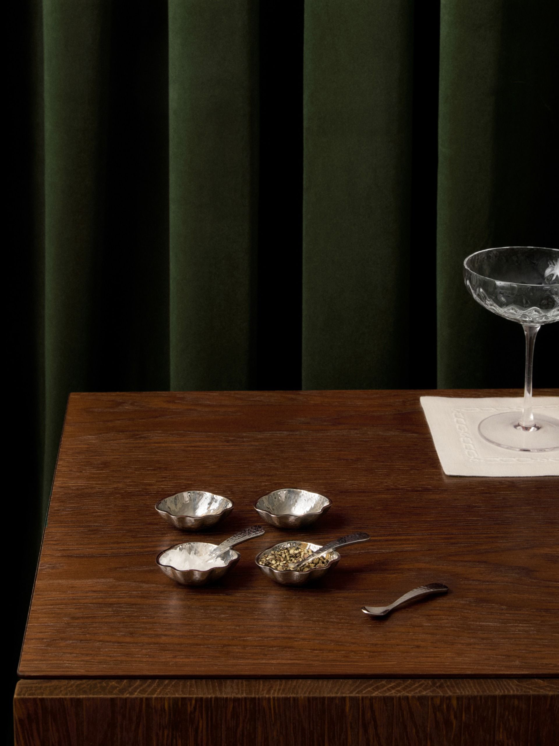 close up of timber table with green velvet curtains in the background, with white cocktail napkin, champagne coupe, and four small salt-and-pepper holders with spoon
