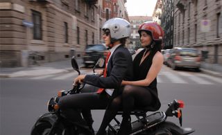 A man with a suit riding a motorbike with a female passenger wearing a red helmet and black dress