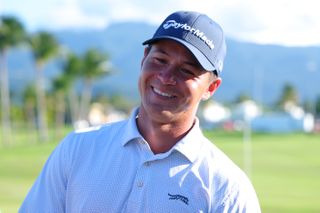 Karl Vilips smiles while wearing a pale Sun Day Red polo and navy blue TaylorMade cap