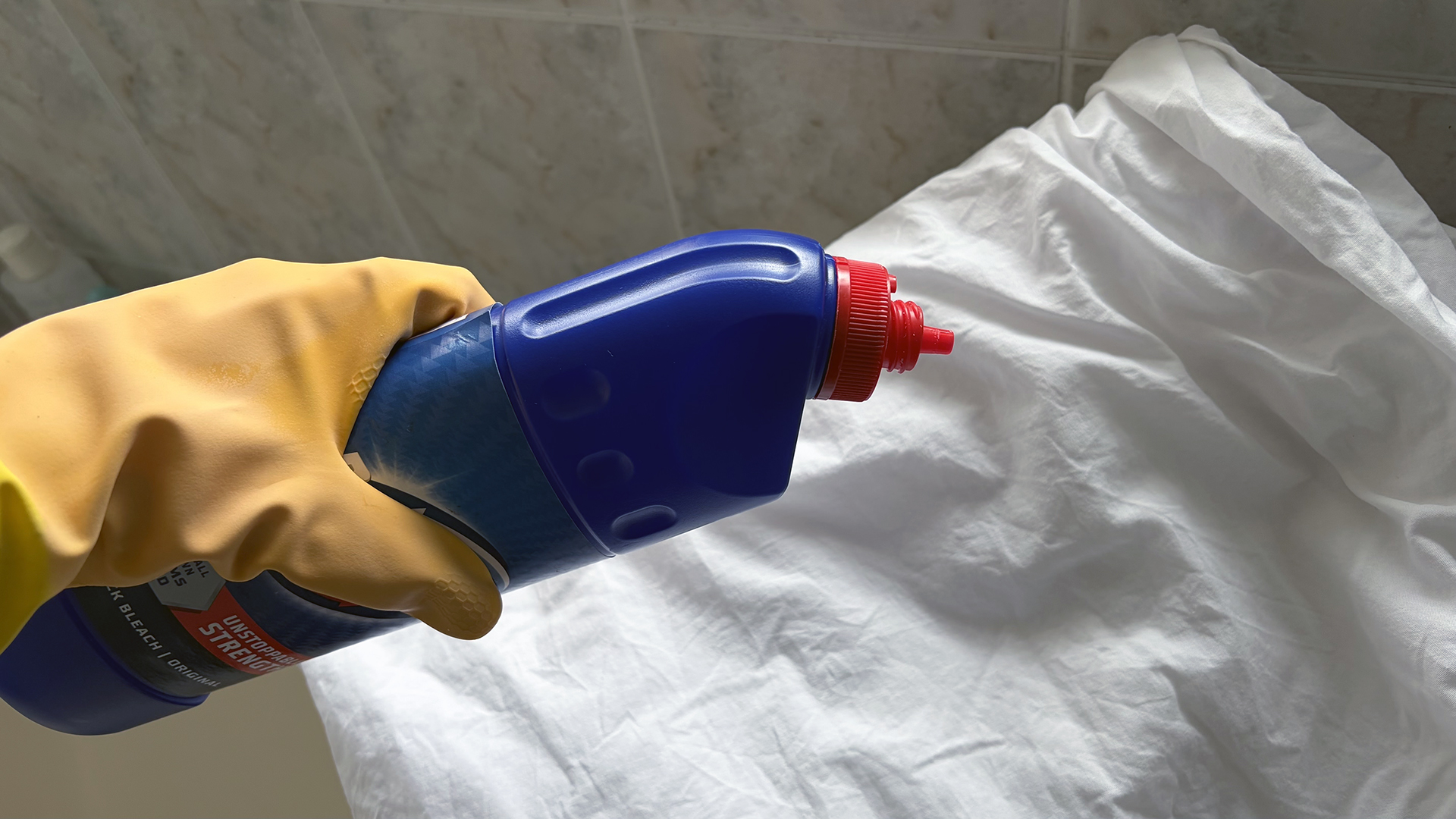 A close up of a hand wearing a rubber glove holding a bottle of chlorine bleach over white bed sheets
