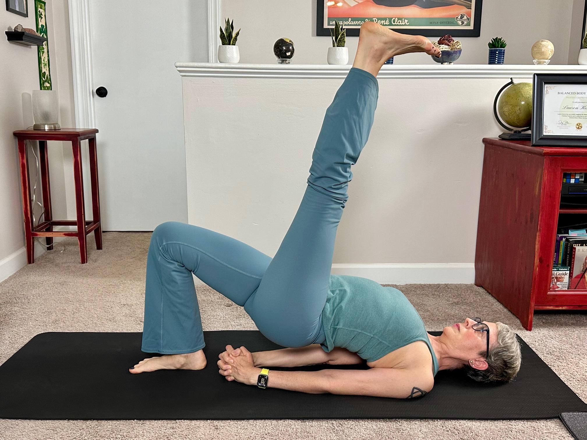 woman performing bridge pose with a single leg on a black yoga mat in a living room