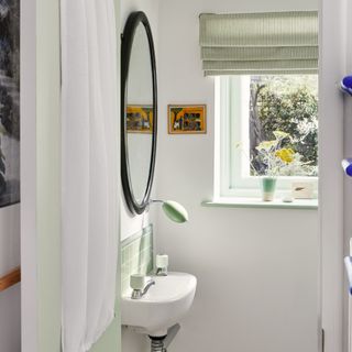 cloakroom design mistakes, white and pale green cloakroom with striped Roman blind, tiny basin, small window with green window sill, black round mirror, green tiled splash back