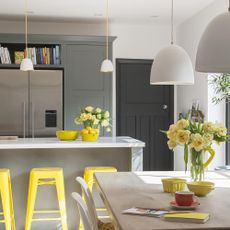 a kitchen and dining area with white walls and a black door, grey cupboards, three yellow stools underneath a breakfast bar, with yellow roses on the table and kitchen bench