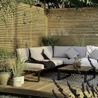 Garden lounge with L shaped sofa, table and potted plants on a wooden deck.