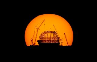 A giant view of the sun, at sunrise, is seen behind a silhouetted ELT under construction.