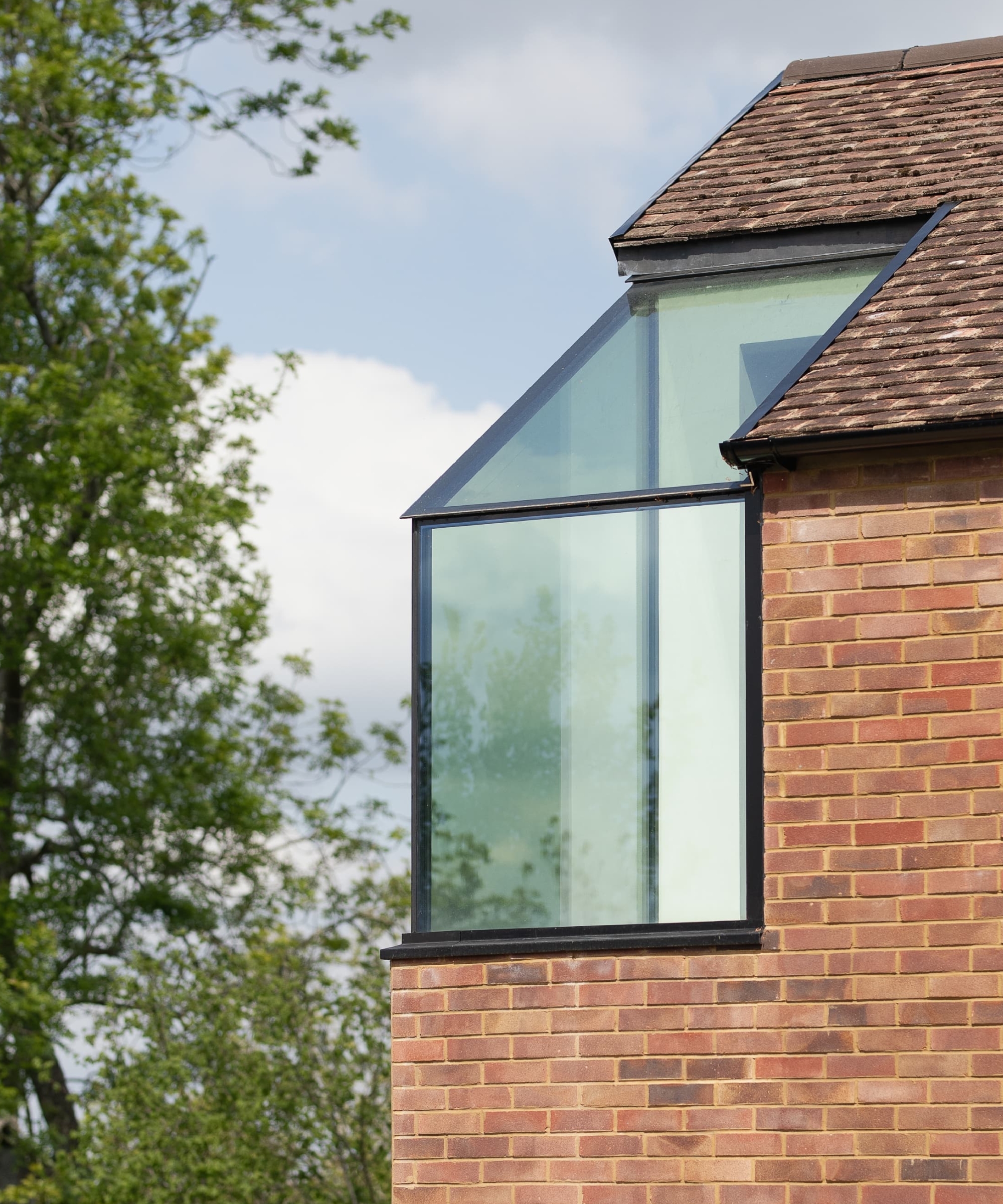 Angled corner window set into a brick house gable, framed by a sloped tiled roof and trees.