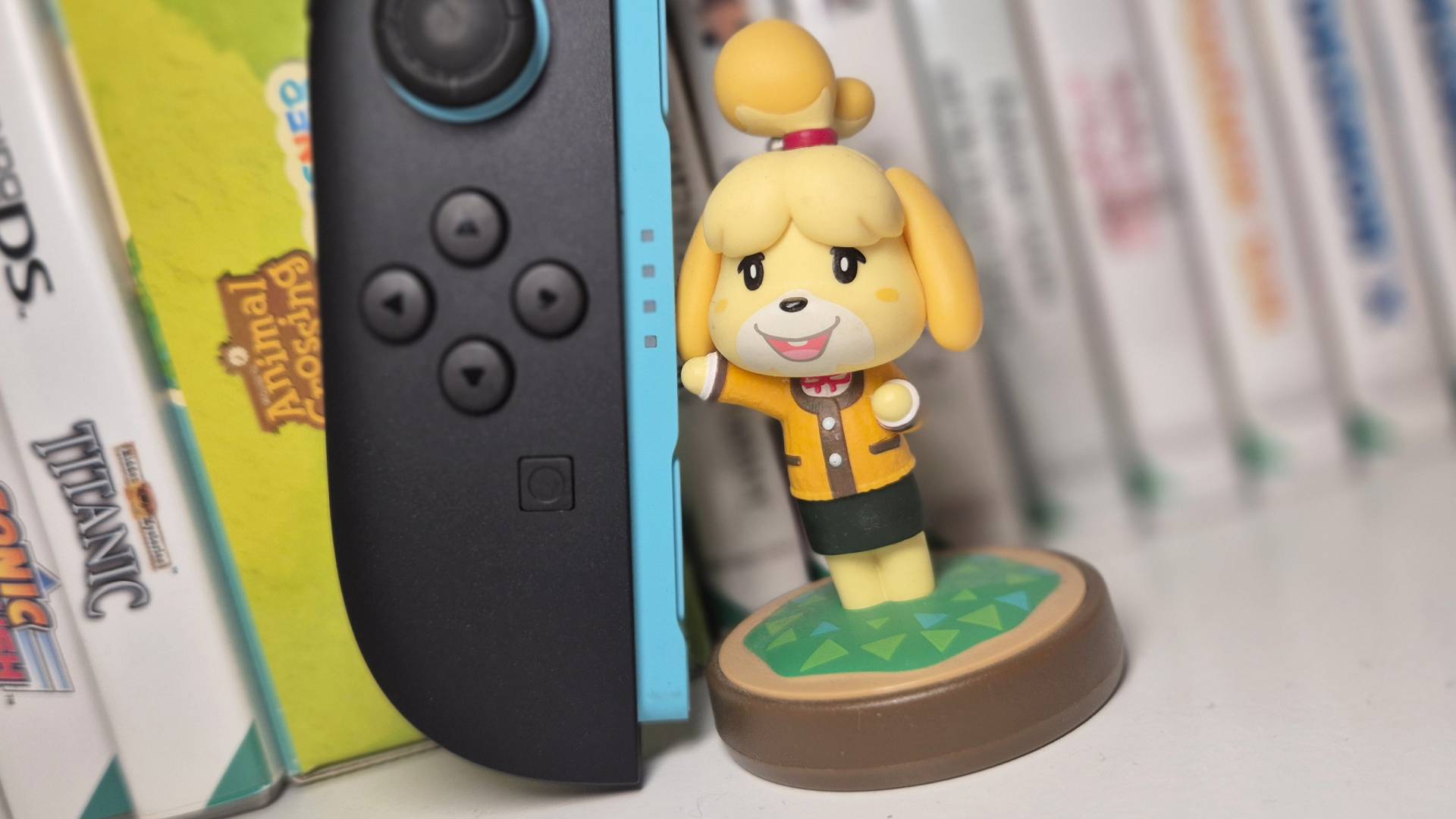 Image of an Isabelle AC amiibo sitting on a game shelf with a left Switch 2 Joy-Con controller.