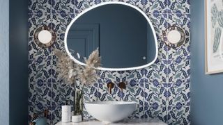 irregular shaped mirror with light around edge on blue floral tiled wall in bathroom