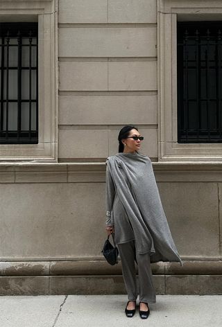 A woman standing outside wearing a gray knitted cape over a gray knit with gray knitted pants, black ballet flats, and a black clutch.