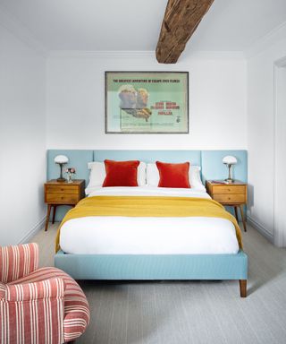 Bedroom with exposed beam, double bed with turquoise oversized headboard and red and white striped armchair on wool carpet.