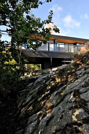 Alternative exterior view of the elevated Trekronekabin - a dark wood house with a chimney and outdoor terrace that sits on rocks surrounded by trees under a blue cloudy sky