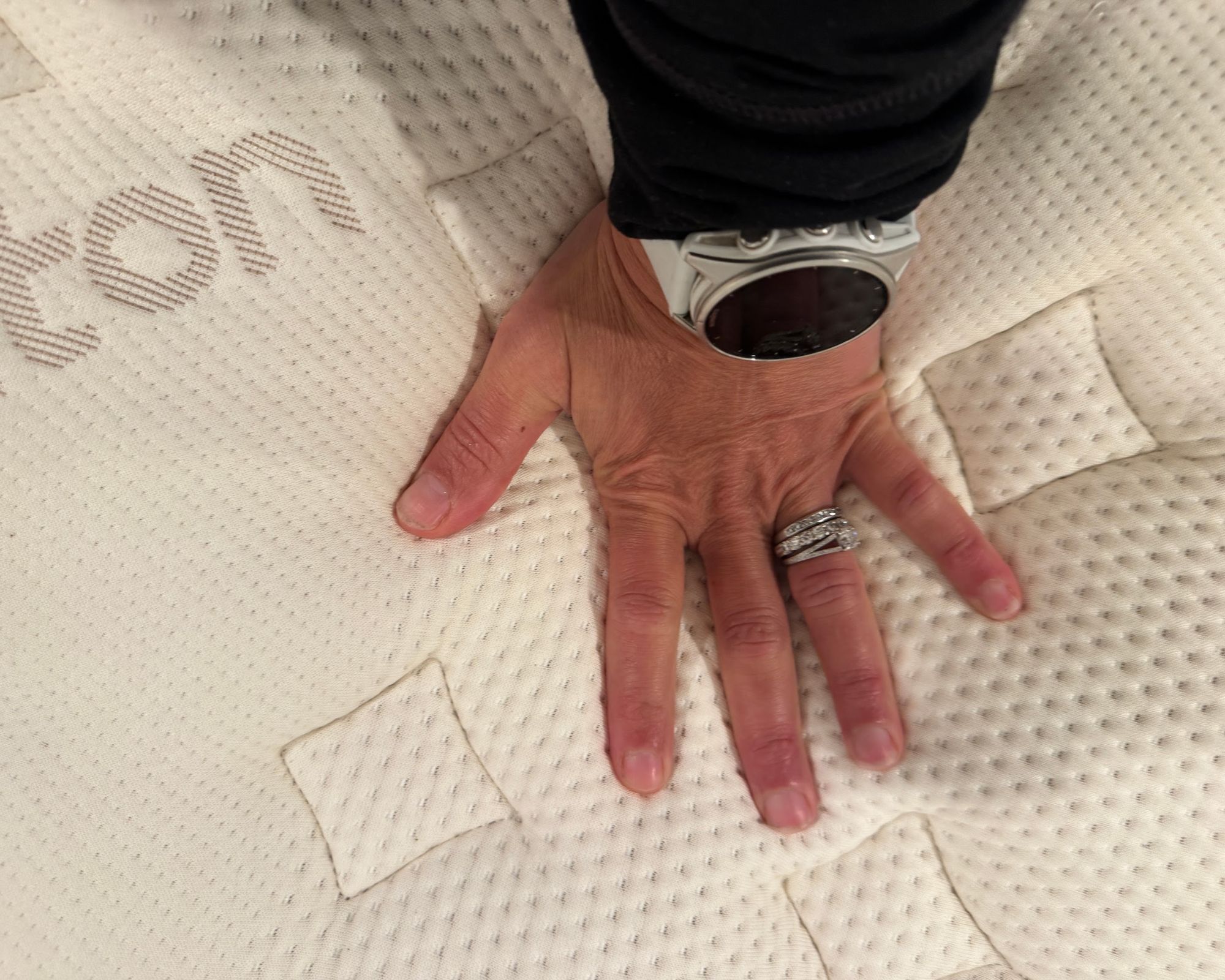 A hand pushing down on a tan mattress. The person pushing on the mattress is wearing silver rings and a watch.