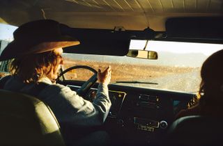 View of Dennis Hopper wearing a cowboy hat and driving a car in the outback. There is another person in the passenger seat next to him