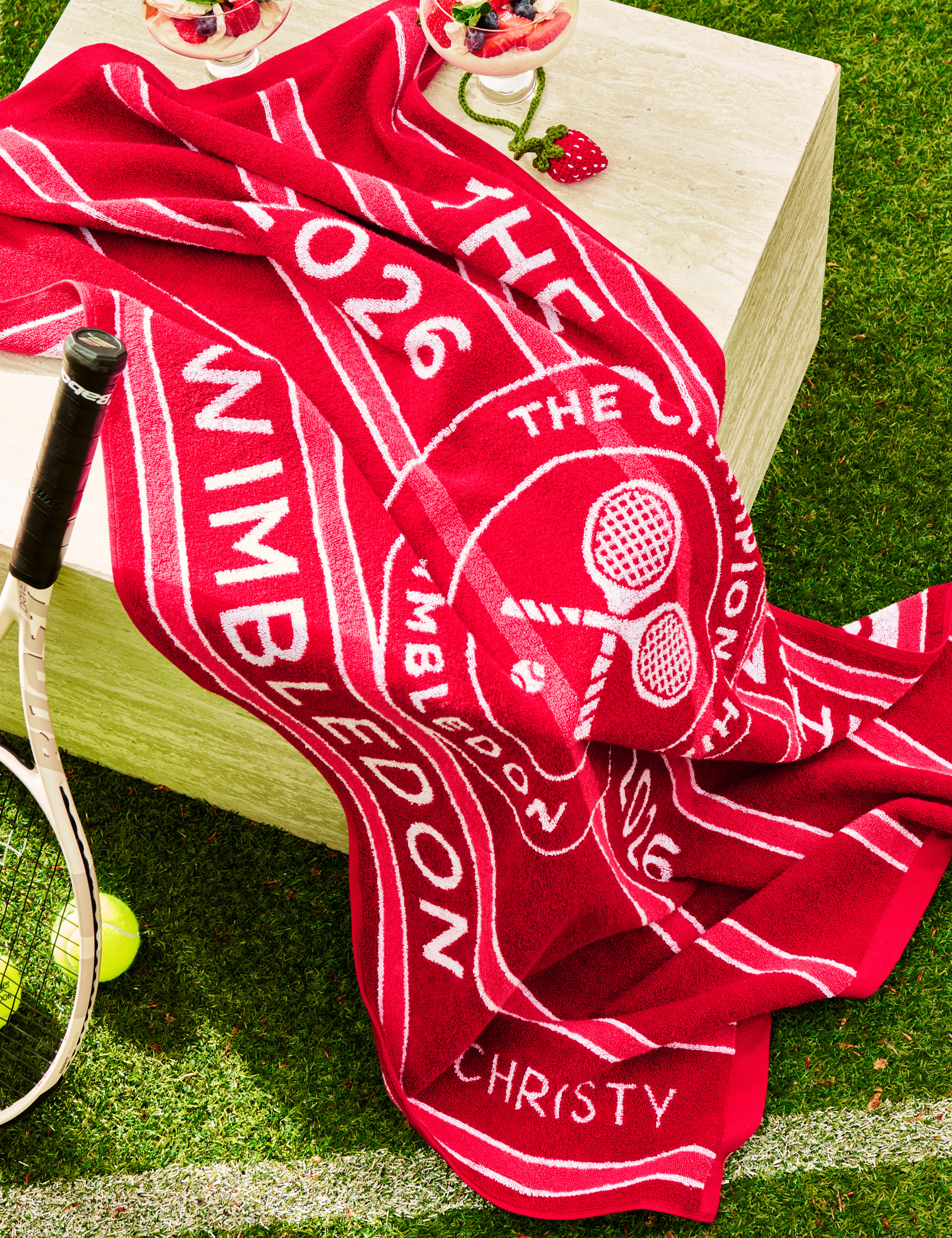 Christy x Wimbledon championship towel pictured in a grassy tennis court alongside a racket and a pair of tennis balls