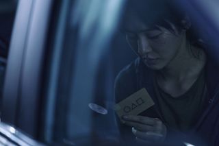 a woman (park gyu-young) looks down at a squid game card that she is holding, while light streams into the room from above