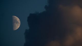 An image of a half-lit moon against a dark blue sky, with its right half lit by direct sunlight and its left shrouded in shadow. Dark plains called lunar maria can be seen covering swathes of the lit side and a large cloud dominates the right side of the image.