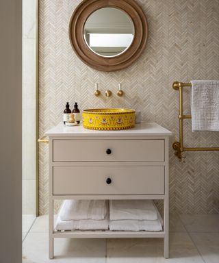 Neutral bathroom with small herringbone wall tiles, round mirror, vanity with yellow patterned basin, brass taps and towel rail, towels in bottom of vanity