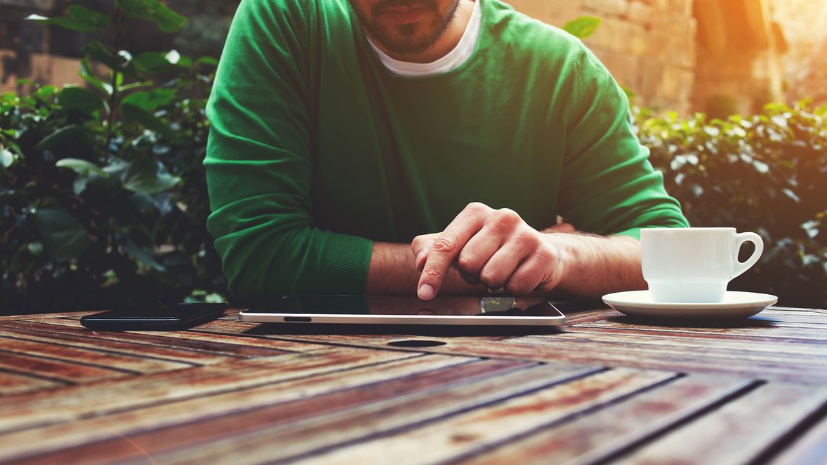 A man sitting at an outdoor table using his tablet with a smartphone next to it on one side and a cup of tea or coffee on the other