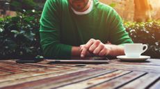 A man sitting at an outdoor table using his tablet with a smartphone next to it on one side and a cup of tea or coffee on the other