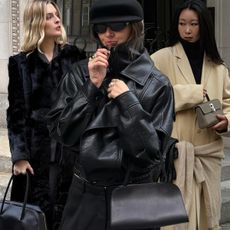 West Village winter trends are showcased in a collage of street-style photos featuring women's winter outfit ideas. On the far left, a woman stands in a black fur coat, holding a black bowler bag. In the middle, a woman is wearing a black leather funnel neck bomber jacket with black trousers, black sunglasses, a black baker cap, and a black bowler bag. On the right stands a woman wearing a black turtleneck sweater layered under a cream coat with a matching cardigan tied around the waist and black gloves layered over the cardigan.