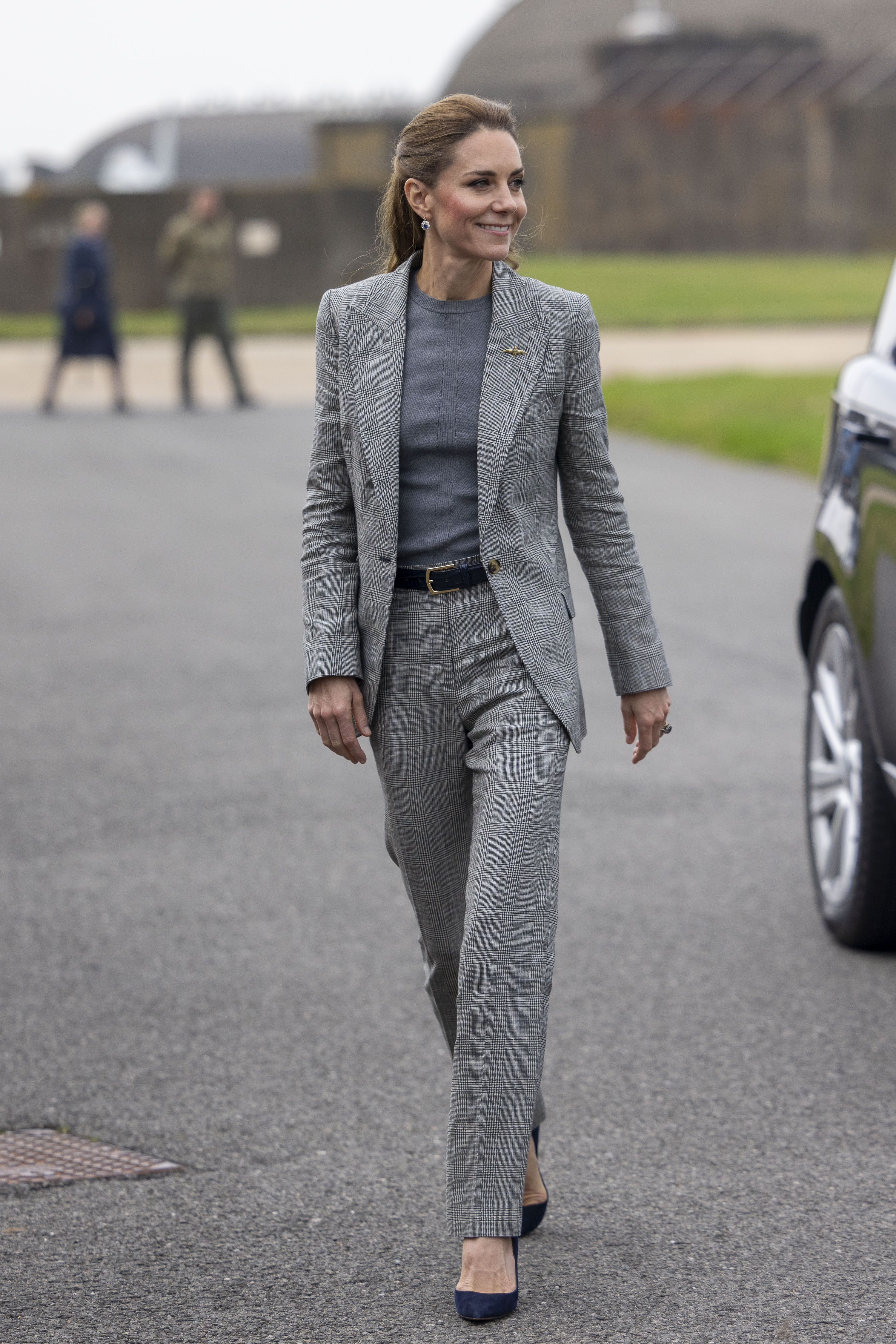 Kate Middleton walks outside wearing a tonal grey suit with navy suede heels and a black leather belt.