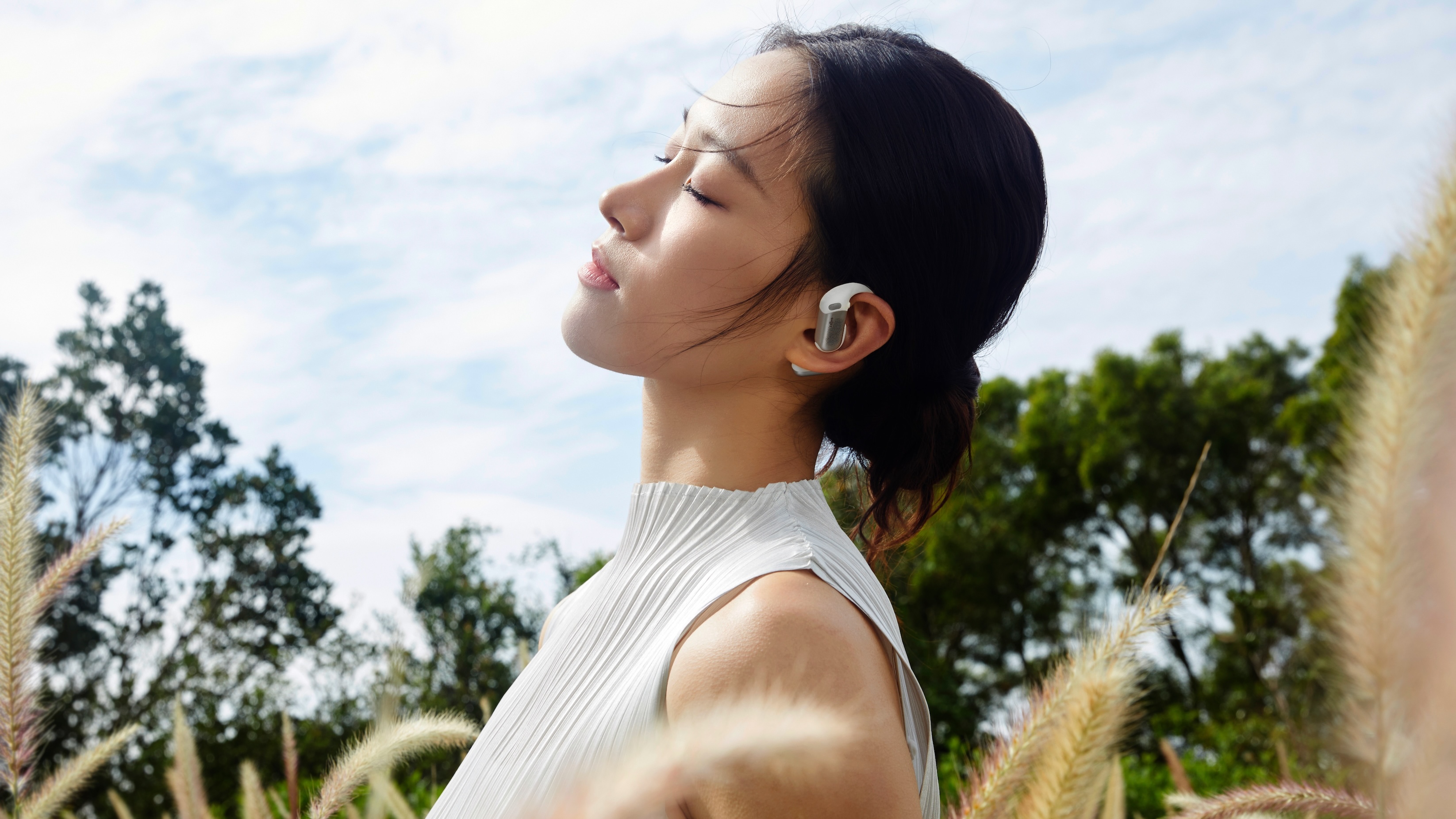 A woman in a field wearing the Shokz OpenFit Pro headphones with her eyes closed.