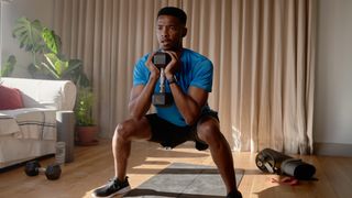 Man performs dumbbell squat at home holding dumbbell close to chest.