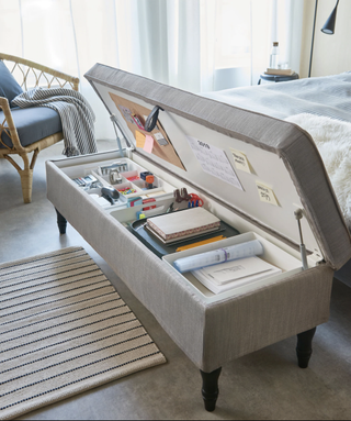 a gray Ikea storage ottoman at the end of a bed, in a neutral bedroom, containing office supplies such as notebooks pens and stationary