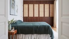 Image of a bedroom with white walls and a floor-to-ceiling built-in, wooden closet. There is a bed with an emerald green minimalist coverlet and a white ruffled bed skirt/valance.