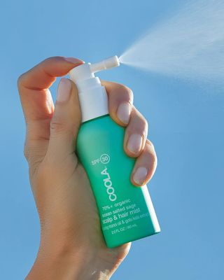 A woman sprays a bottle of Coola Scalp & Hair Sunscreen Mist