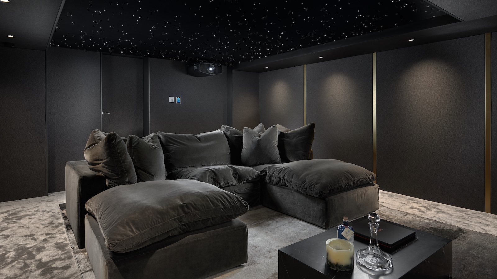 A photograph of a luxury, custom-installed home cinema with a huge projector screen, lush u-shaped sofa, grey walls and lamps