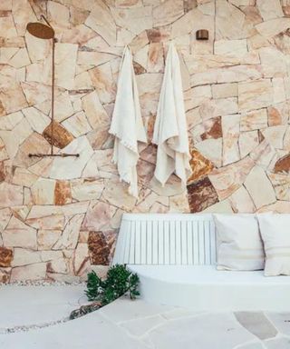 An outdoor shower area with a copper shower head, a light pink stone wall with towels hung on it, and a white long outdoor seat