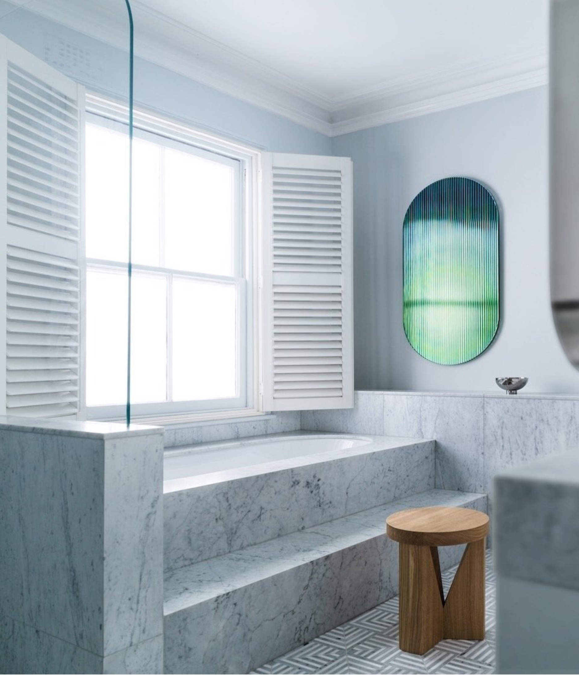 a white bathroom with a step up bath and green and blue ombre mirror and wooden stool