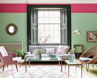 Bright, colorful living room with green painted walls and pink painted trim, large window with wooden shutters, light wooden flooring, cream and pink rug, gray sofa, pink armchair, wicker style armchair, wooden and glass coffee table, artwork and mirrors on wall
