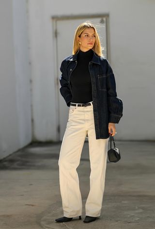 A cute work winter outfit is shown in a photo of a woman standing outside wearing a dark wash denim jacket over a black turtleneck with a black belt, white wide-leg jeans, black ankle boots, and a black handbag