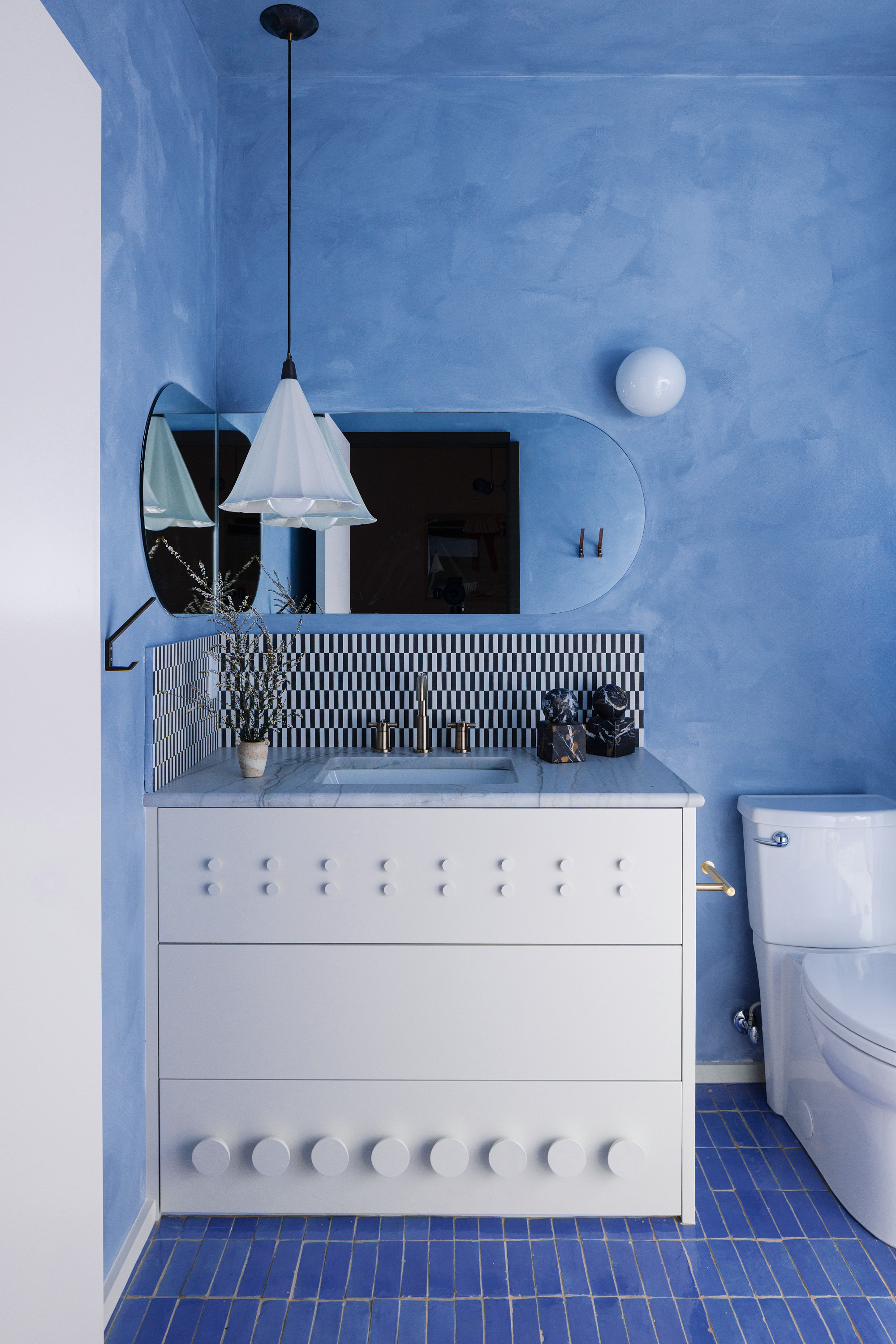 Bathroom with blue limewash walls, blue tiled floor, white vanity with black and white checkerboard backsplash tiles, and rounded corner mirror