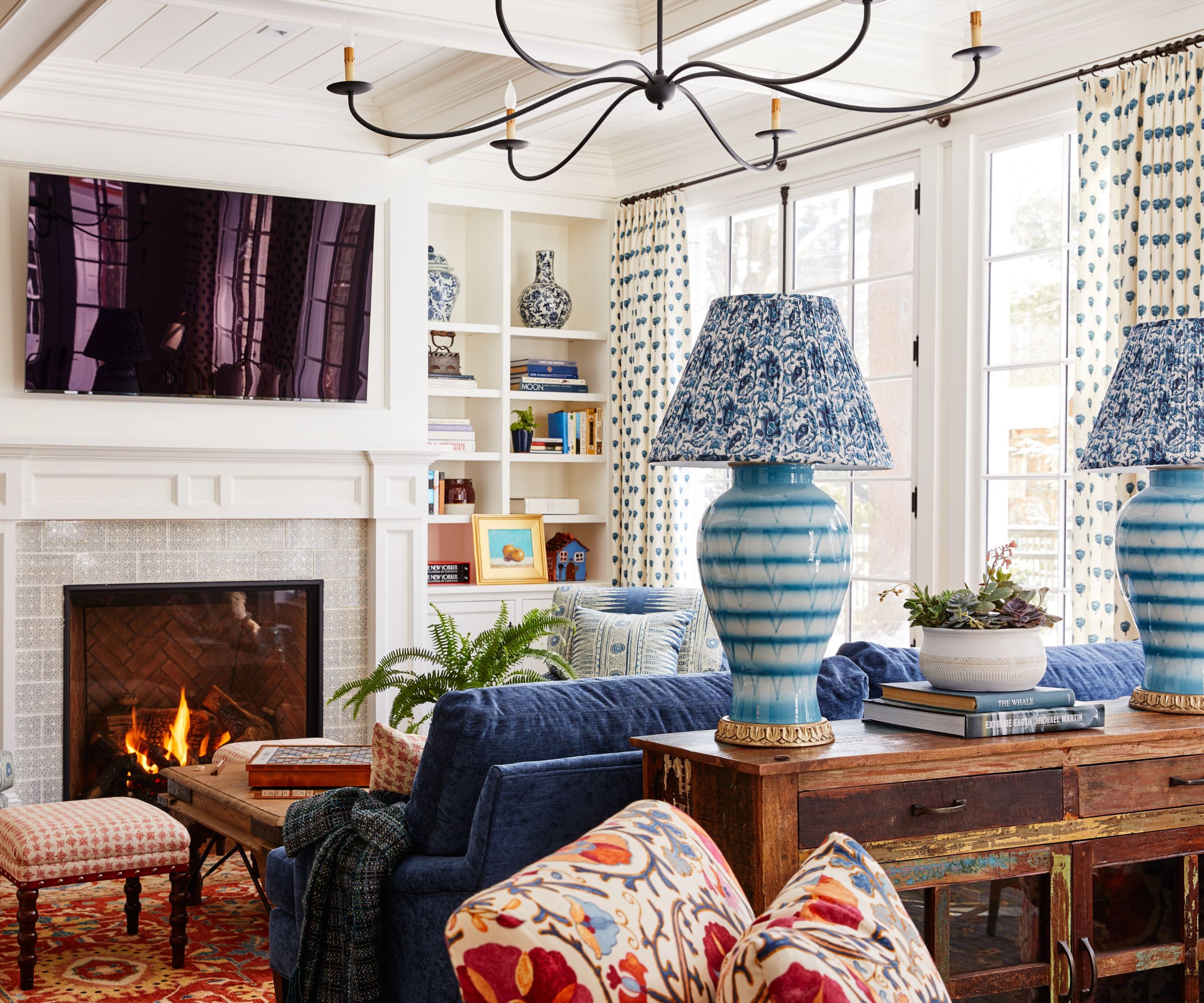 Eclectic living room with Persian rug, blue sofa, wooden console with blue table lamps on either side, floral armchair, large fireplace with TV above, and built-in bookshelves with books and decor