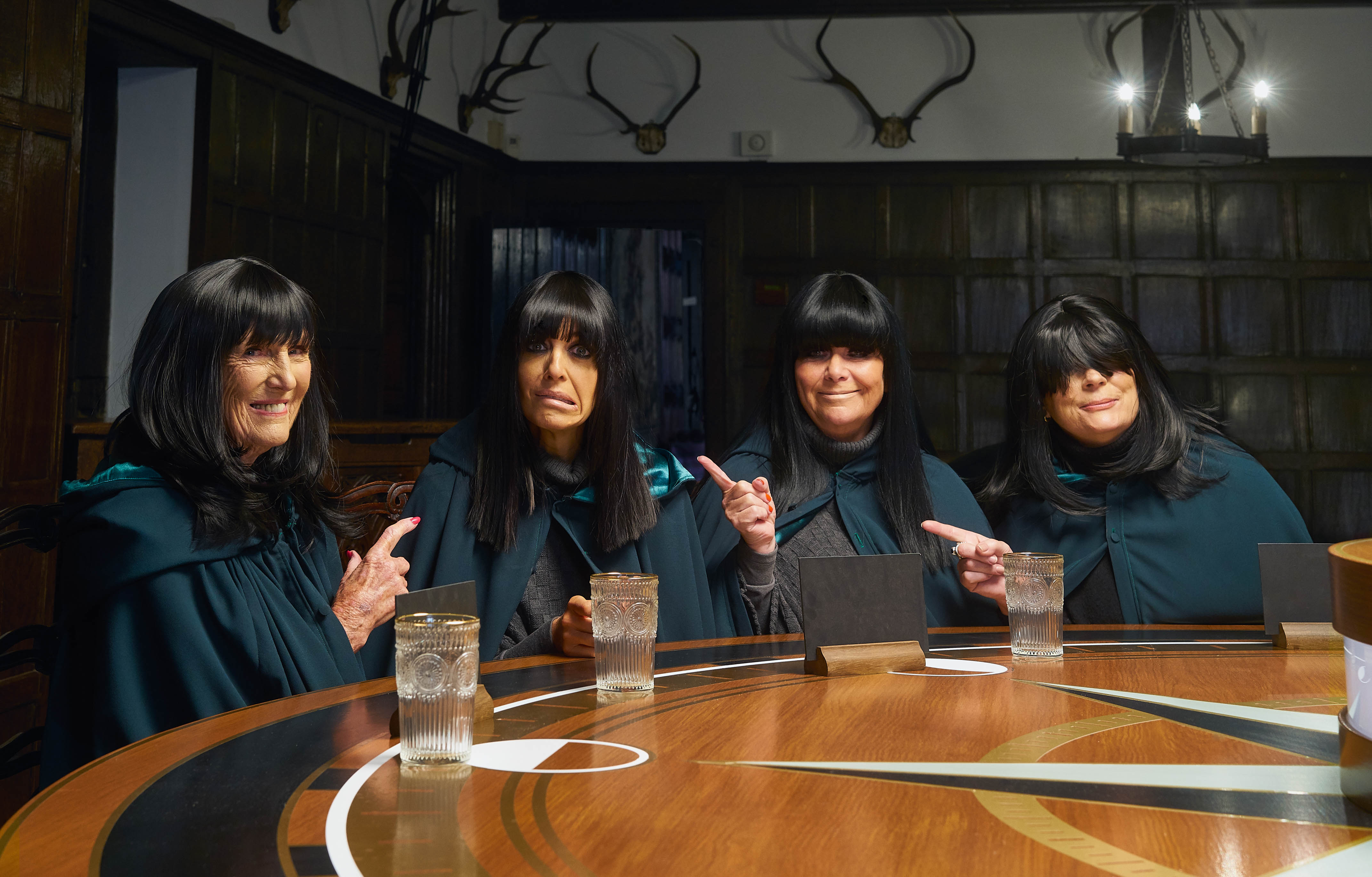 Mary Berry, Claudia Winkleman, Dawn French and Jennifer Saunders sit around The Traitors roundtable. All of them are wearing black Traitor cloaks, and Mary, Dawn and Jennifer are all wearing black wigs with long fringes mimicking Claudia's hairstyle