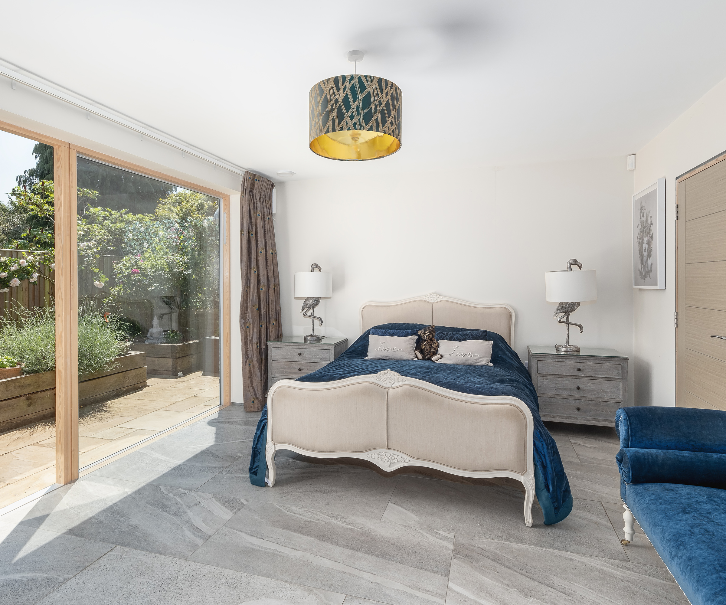 A ground-floor bedroom with pale grey tiled floor and a large antique-style bed. There is floor-to-ceiling glazing with a sliding glass door leading outside
